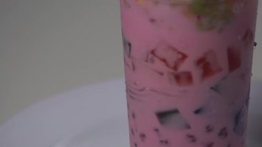 Beyaz bir tabakta dönen plastik bir bardakta ferahlatıcı bir Endonezya buzlu tatlısının aşırı yakın çekimi. Renkli jöle küpleri, fesleğen tohumları ve meyve parçalarıyla karıştırılmış canlı pembe süt şurubu. Yaz içkileri ve Asya mutfağı için mükemmel.