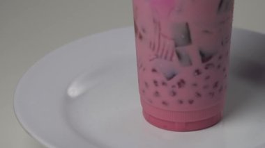 Beyaz bir tabakta plastik bir bardakta çimen jölesi ve fesleğen tohumu ile taze pembe süt içeceği. Popüler Güneydoğu Asya sokak yemekleri, yaz içeceği, Ramazan iftarı veya kafe menüsü sergisi için mükemmel. 4K yüksek kaliteli görüntü.