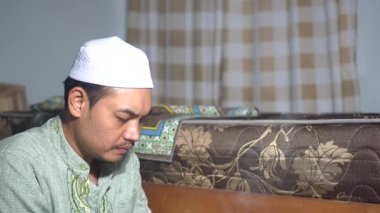 Güneydoğu Asyalı, beyaz şapkalı Müslüman bir adamın evde sessizce dua edişinin 4K görüntüsü. Kapalı mekan aydınlatması dini, Ramazan veya Kurban Bayramı temalı içerik projeleri için barışçıl ve ruhani bir ortam yaratır..