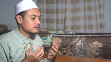 Asyalı Müslüman bir adam, beyaz bir dua şapkası ve geleneksel yeşil bir gömlek giyip evinde kutsal bir dua ediyor. Sadakatle ellerini kaldırır ve yüz silme ile bitirir. Ruhani, barışçıl İslami yaşam tarzı ve ibadet kavramı.