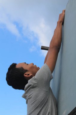 Gri tişörtlü, gri bir duvara tırmanan ya da açık mavi bir gökyüzüne asılan genç bir adamın yan profili. Çaba, mücadele, hırs ve hedeflere ulaşma kavramı. Dinamik düşük açı çekimi güç ve kararlılık gösteriyor