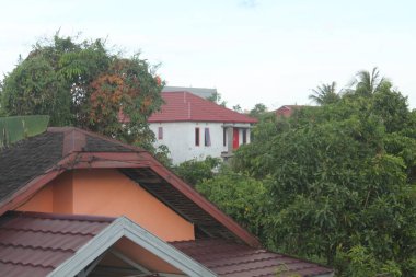 Bereketli tropik yeşilliklerin arasına yerleştirilmiş kırmızı kil kiremit çatılı evleri yüksek açılı görüyor. Parlak bir öğleden sonra gökyüzü altında kentsel gelişim ve doğa arasındaki güzel tezat.