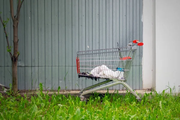 An abandoned cart from a store with garbage inside stands on the grass against a gray metal fence