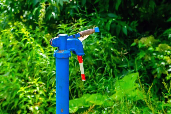 The hand water pump of metal is painted blue. Green. Street. Outdoor.