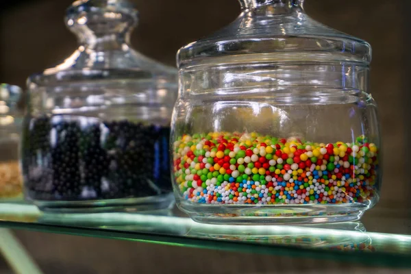 Glass jar with multi-colored round topping for ice cream. Stands on a glass display case.