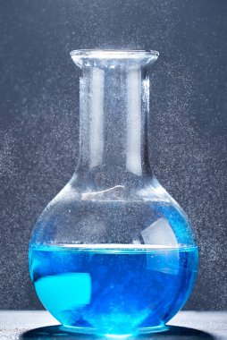 Gri zemin üzerinde mavi bir sıvı bulunan cam şişe..