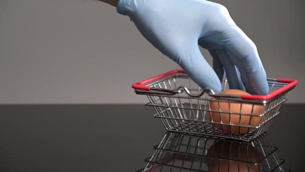 Mavi eldivenli bir el, yumurtaları minyatür bir alışveriş sepetine koyar. Güvenli alışveriş konsepti. — Stok video