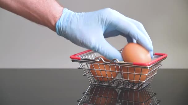 Uma mão em luva médica azul remove ovos de uma cesta de compras em miniatura. Conceito de compra segura. — Vídeo de Stock