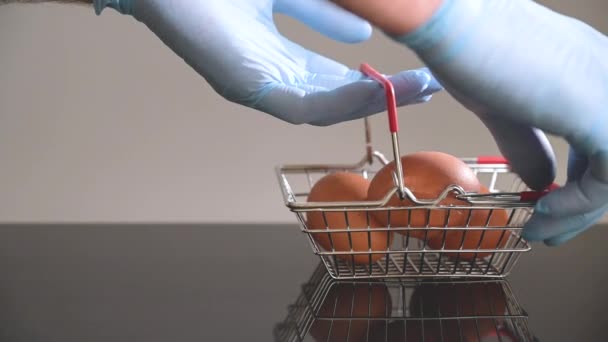 Een hand in de blauwe medische handschoen pakt een miniatuur boodschappenmandje met eieren. Veilig winkelen concept. — Stockvideo