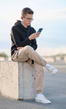 Kapüşonlu, sıradan pantolonlu ve beyaz ayakkabılı şık bir genç. Kulaklıklı bir adam oturuyor ve gökyüzüne karşı akıllı telefon kullanıyor..