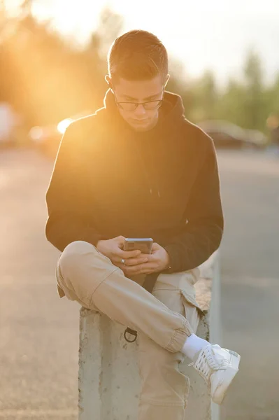 Un joven con estilo en una sudadera con capucha, pantalones casuales y zapatillas blancas. Un tipo con auriculares está sentado y usando un teléfono inteligente. Foto tonificada naranja. — Foto de Stock