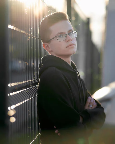 Porträt eines jungen, stylischen Typen mit Brille. Ein ernsthafter junger Mann steht neben einem eisernen Zaun. Sonnenlicht fällt von hinten. — Stockfoto