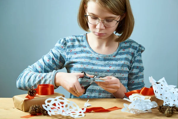 Girl teenager with glasses carving snowflakes out of paper. Concept preparation for the New year. Royalty Free Stock Photos