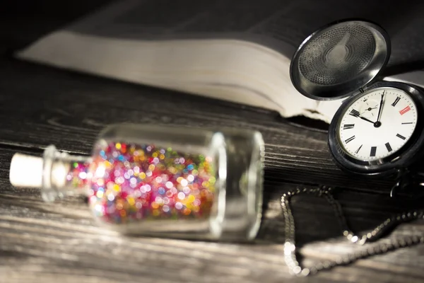 Una pequeña botella de vidrio con brillantes cuentas de colores brillantes y tapa de corcho se encuentra junto al viejo reloj de bolsillo y un libro abierto iluminado por una bombilla. Foto en secreto. Concéntrate en reloj —  Fotos de Stock