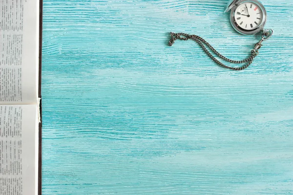 Винтажные серебряные карманные часы и открытая книга лежат на углу потрепанного старого синего деревянного фона. Вид сверху — стоковое фото