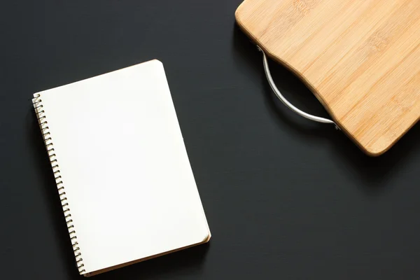 Old empty wooden cropped cutting board and notepad with blank for picture or inscription on black background from chalkboard. Close up. Top view. Flat lay. Mock up for restaurant menu.