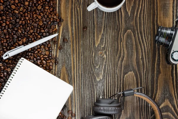 Cup of coffee, headphones for music single simple empty white notebook with a blank for drawing or writing, white plastic pen and scattered roasted coffee beans are on a Desktop from wooden plank. Top