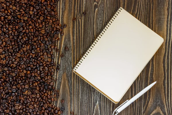 Ein einzelnes leeres weißes Notizbuch mit einem Rohling zum Zeichnen oder Schreiben, weißem Plastikstift und verstreut gerösteten Kaffeebohnen liegt auf einem Schreibtisch aus Holzplanken. Ansicht von oben. Attrappe. flache Lage — Stockfoto