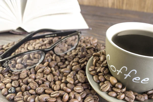 Cup of coffee  with beans in front of open book  standing on a wooden table.  Crop, blurred, focus on mug — Stock Photo, Image