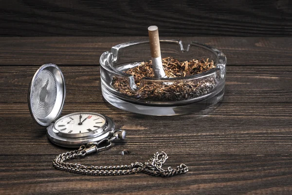 古董怀表和皱巴巴的香烟，在旧木桌上的玻璃烟灰缸 — 图库照片