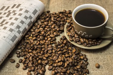 Bir fincan kahve dağınık kahve çekirdekleri ile standları yanındaki bir dergi bulmaca bir parça çuval bezi vardır