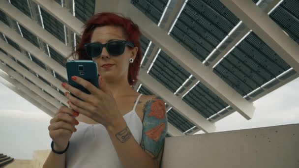 Mujer tatuada usando su teléfono inteligente con un gran panel solar 4k UHD disparo — Vídeos de Stock