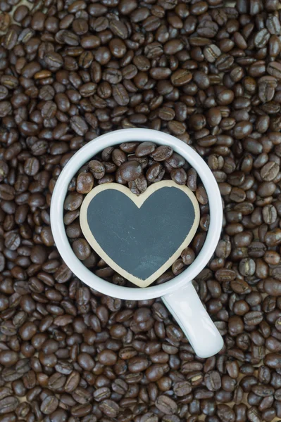 Caneca branca e forma de coração de madeira no fundo de grãos de café — Fotografia de Stock