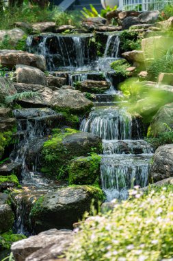 Yosun kaplı kayaların ve yemyeşil çimlerin arasından akan ferahlatıcı bahçe şelalesi. Doğal taşlar ve akan su içeren sakin ve huzurlu bir bahçe tasarımı..