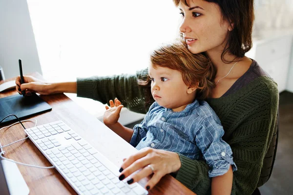 Cropped shot of a young woman holding her son while doing some work on her computer