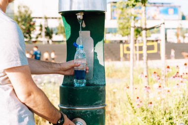 Şehir parkında halka açık bir çeşmede plastik su şişesini dolduran adam..
