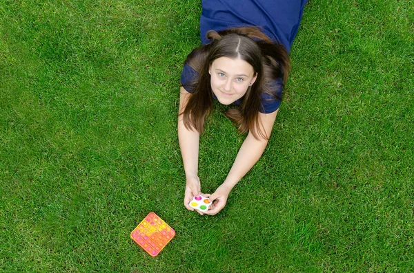 A teenage girl is playing with simple dimple and popit anti stress toys outside on the lawn, looking up at the camera.