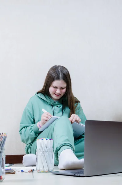Egy fehér tinédzser lány ül a padlón a laptop előtt, és rajzol a jegyzetfüzetére.. — Stock Fotó