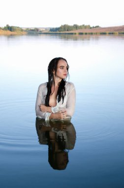 Beyaz tişörtlü, hayalperest genç bir kadın gölde duruyor, kollarını sarıp, başka tarafa bakıyor..