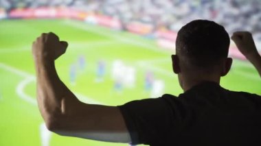 Amigo bir adam arka planda bir spor maçı izliyor. Futbol taraftarı iç sahadaki takımı için tezahürat yapıyor. Spor etkinlikleri görüntüleniyor