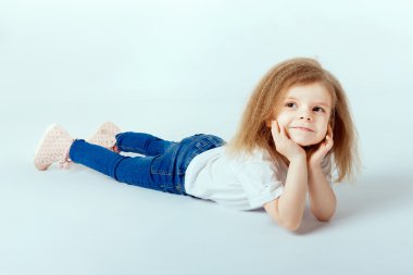 little girl 4 years old with curly hair wearing white shirt, blue jeans lying on the floor, smiling and looking at camera, hands holding her head