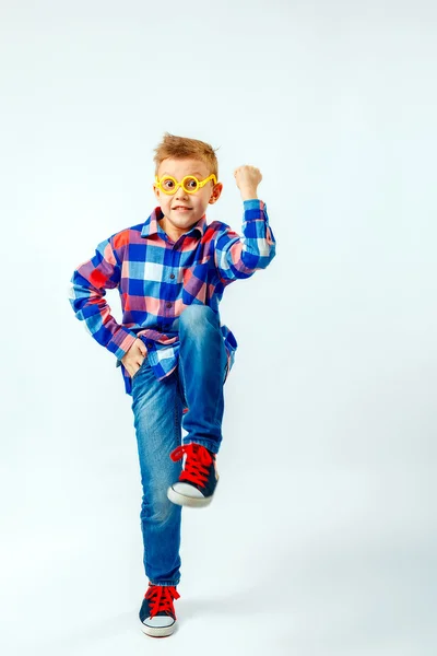 Little boy wearing colorful plaid shirt, blue jeans, gumshoes, plastic glasses having fun in the studio — Stockfoto