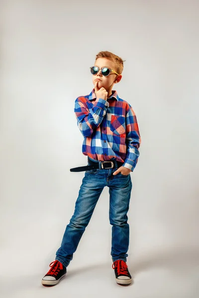 Niño con camisa a cuadros de colores, vaqueros azules, zapatos de goma, gafas de sol, posando y divirtiéndose en el estudio — Foto de Stock