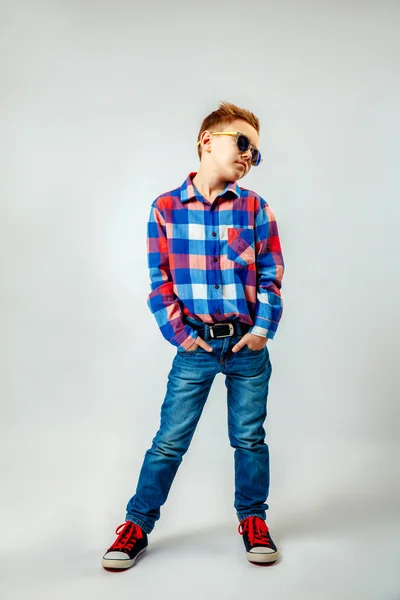 Niño con camisa a cuadros de colores, vaqueros azules, zapatos de goma, gafas de sol, posando y divirtiéndose en el estudio — Foto de Stock