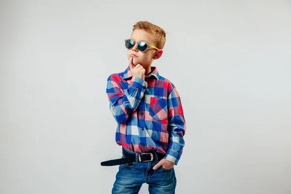 Niño con camisa a cuadros de colores, vaqueros azules, zapatos de goma, gafas de sol, posando y divirtiéndose en el estudio — Foto de Stock