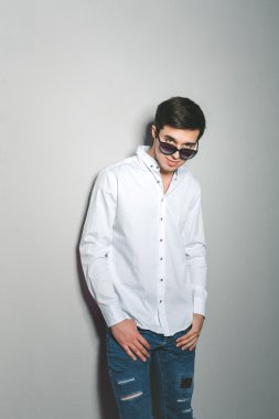 Şortlu ve beyaz gömlekli genç adam gülümsüyor. Gözlüklü duvarın yanında duruyor.