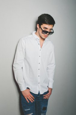 Şortlu ve beyaz gömlekli genç adam gülümsüyor. Gözlüklü duvarın yanında duruyor.