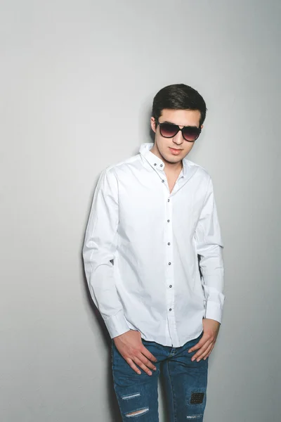 Jovem de shorts e camisa branca está sorrindo de pé perto da parede com óculos — Fotografia de Stock