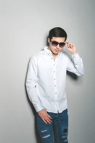 Hombre joven en pantalones cortos y camisa blanca sonríe de pie cerca de la pared con gafas —  Fotos de Stock