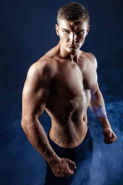 Hezký moc atletický muž kulturista. Fitness svalnaté tělo na tmavé kouřové pozadí. Dokonalý muž. Úžasné kulturista, tetování, pózuje. — Stock fotografie