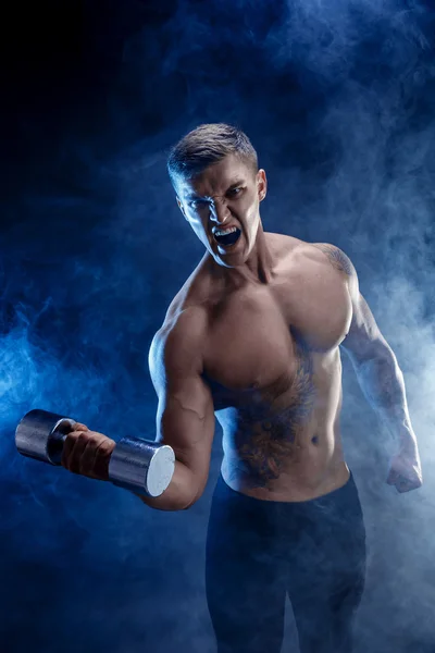 Closeup bir yakışıklı güç atletik erkek vücut dumbbell ile yapıyor. Fitness kas vücut koyu duman arka plan üzerinde. Mükemmel erkek. Harika vücut geliştirmeci, dövme, poz. — Stok fotoğraf