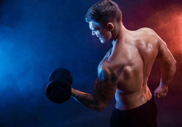 Primer plano de un atractivo poder atlético hombre culturista haciendo ejercicios con mancuerna. Cuerpo muscular fitness sobre fondo de humo oscuro. Hombre perfecto. Culturista impresionante, tatuaje, posando . — Foto de Stock