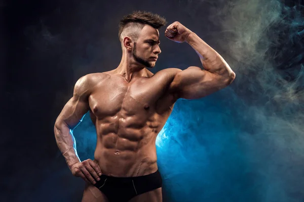 Όμορφος δύναμη αθλητική άντρας bodybuilder. Υγρή Fitness μυώδες σώμα σε σκούρο φόντο καπνού. Τέλειο αρσενικό. Φοβερό bodybuilder, θέτοντας. — Φωτογραφία Αρχείου