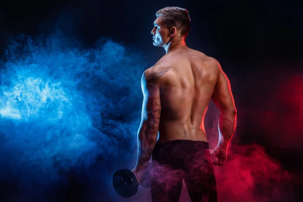 Närbild av en stilig power atletisk man bodybuilder vilar medan stativ med hantel. Konditionen muskulös kropp på mörk rök bakgrund. Perfekta hane. Awesome bodybuilder, tatuering, poserar. — Stockfoto