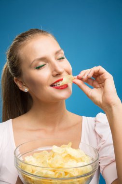 Pembe-kırmızı elbise, güzel dudaklar, holding, kızarmış patates, patates kızartması, cips yeme ve mavi arka plan üzerinde poz parlak makyaj moda genç sarışın kadın portresi. Sağlıksız yeme. Abur cubur
