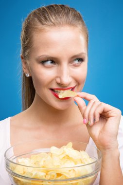 Pembe-kırmızı elbise, güzel dudaklar, holding, kızarmış patates, patates kızartması, cips yeme ve mavi arka plan üzerinde poz parlak makyaj moda genç sarışın kadın portresi. Sağlıksız yeme. Abur cubur